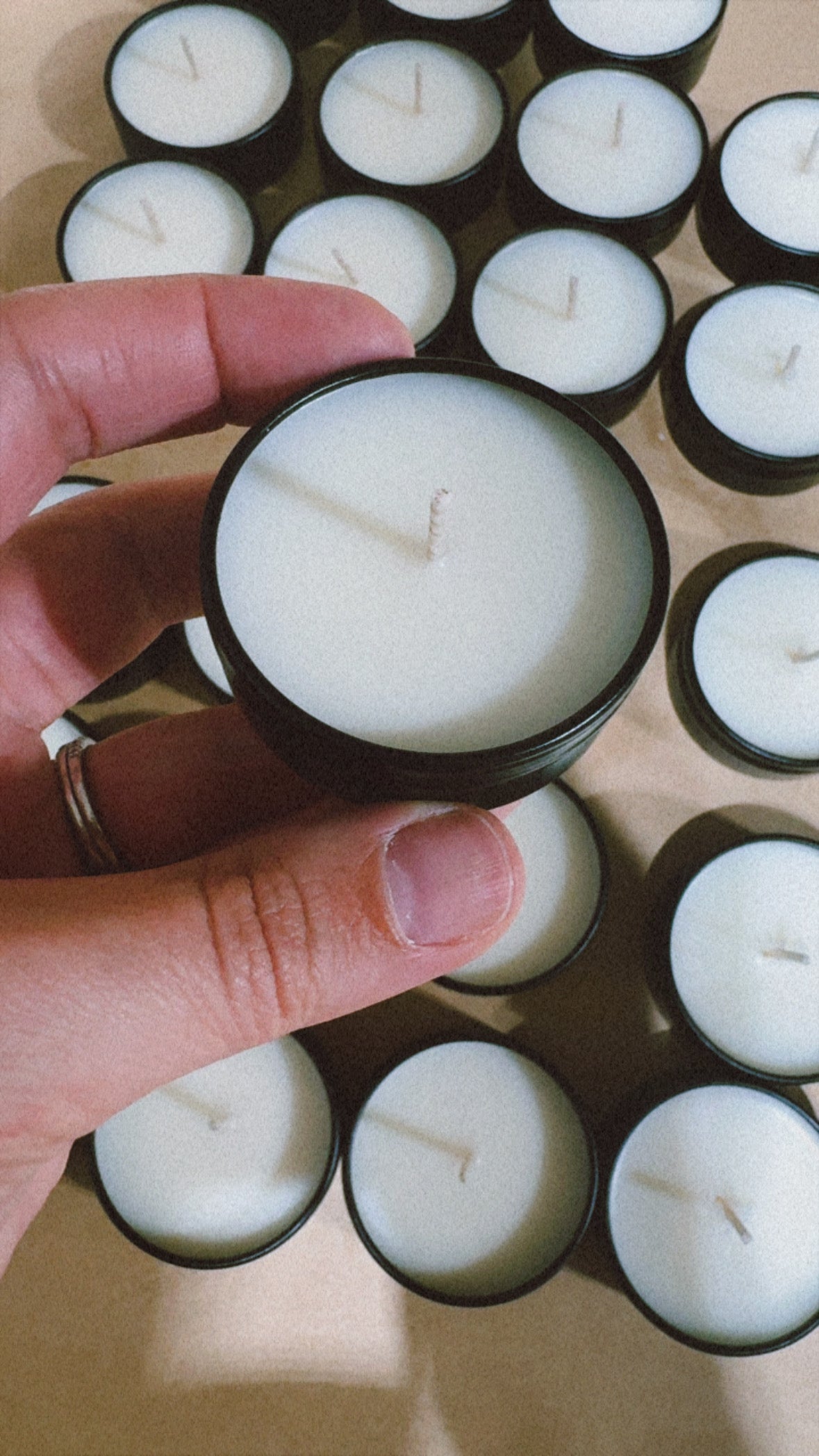 A hand holding a small black tin candle with white wax, with many similar candles arranged in the background on a light surface.