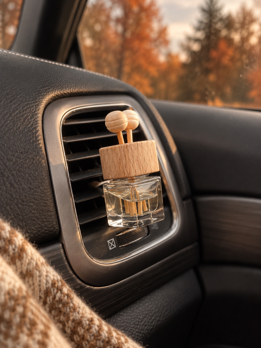 A car air freshener with a wooden diffuser cap and reeds, clipped onto a car dashboard air vent. The background shows a blurred car interior and autumn trees.