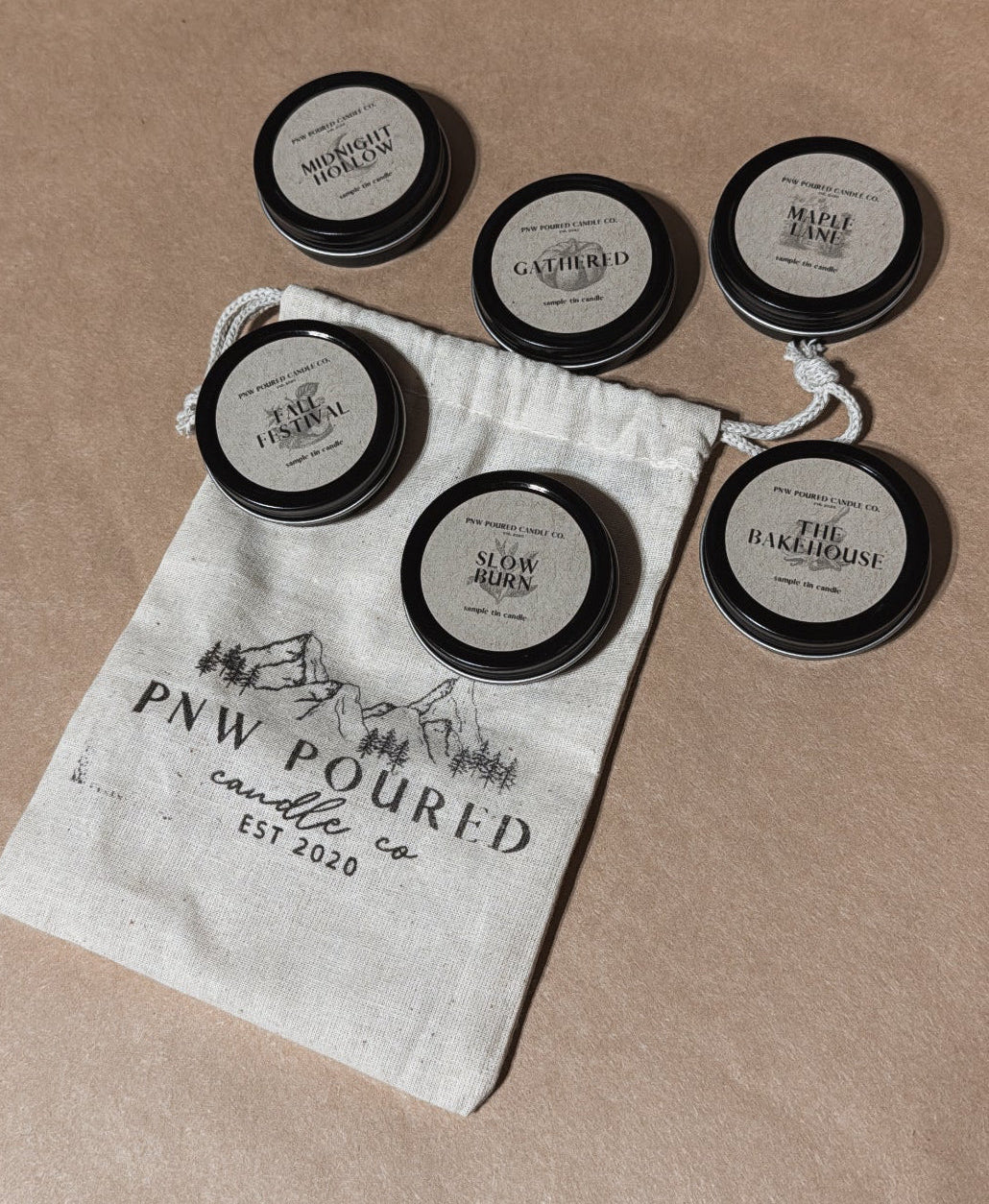 Six small round black-lidded sample tins with kraft brown labels are arranged on a beige surface next to a white drawstring pouch printed with the brand name PNW Poured.